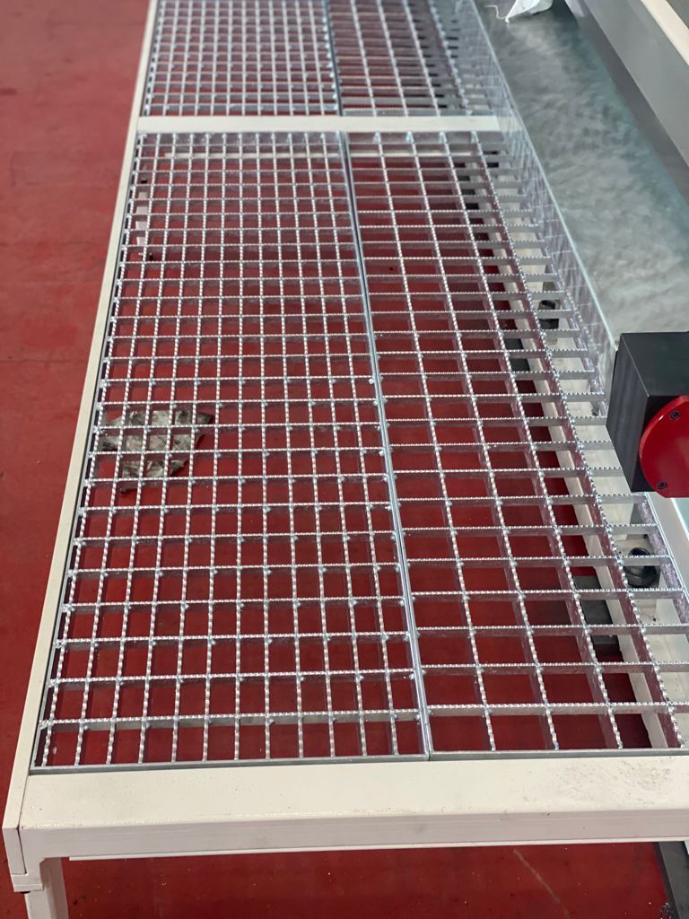 Passerella in grigliato metallico con telaio bianco e superficie inferiore rossa, accanto a un marciapiede e a un semaforo rosso.