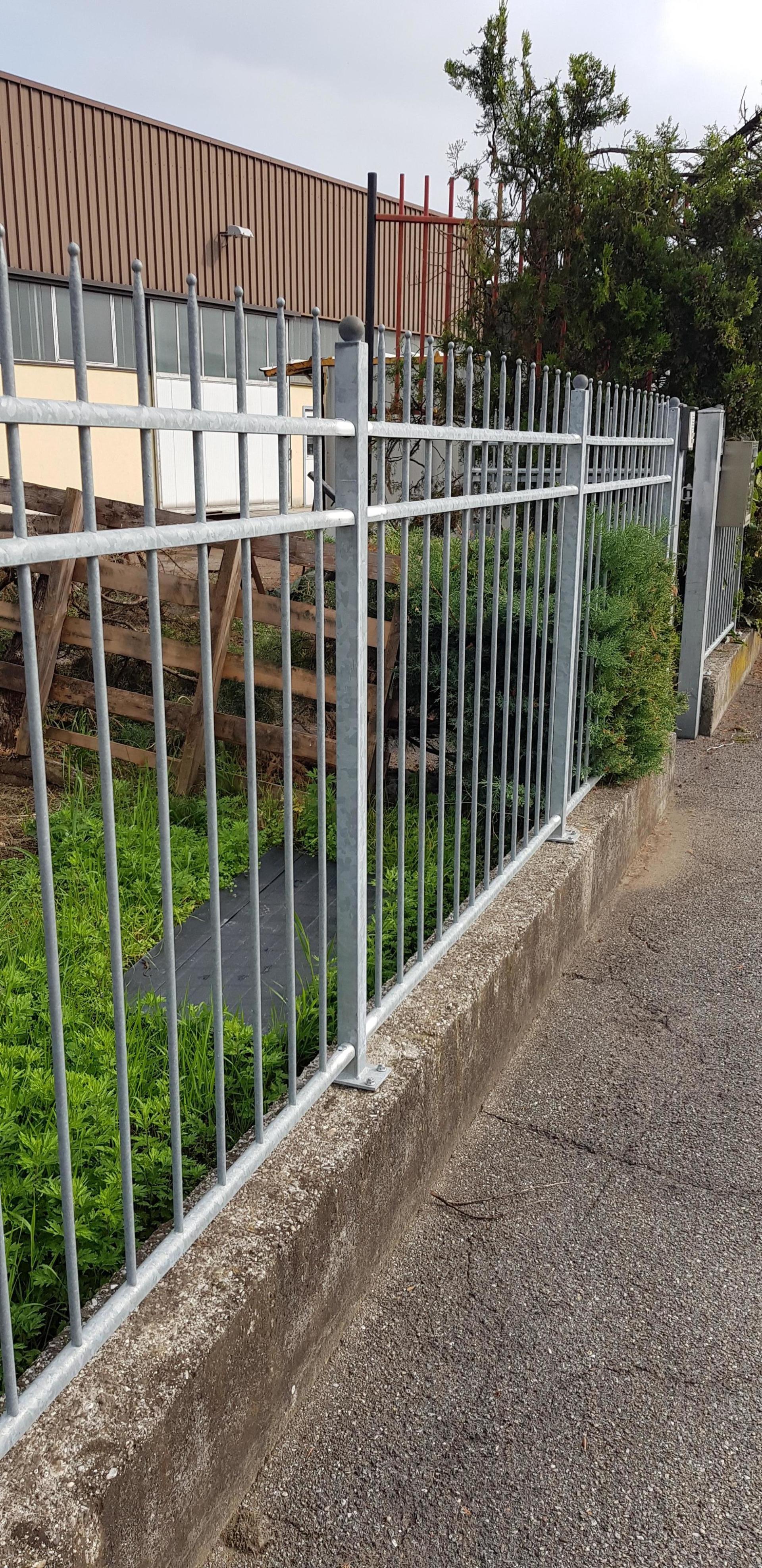 Recinzione metallica bianca lungo un sentiero di ghiaia accanto a un edificio marrone e alla vegetazione.