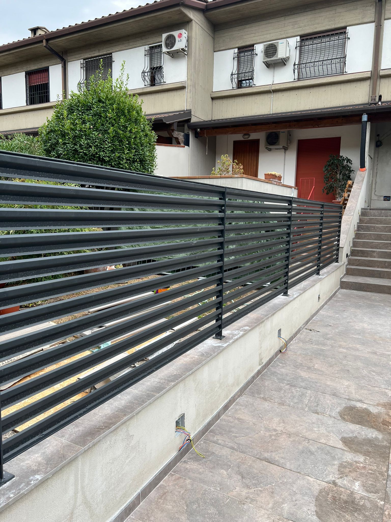 Ingresso di un condominio con porta rossa, scala e recinzione metallica scura lungo un vialetto di cemento.
