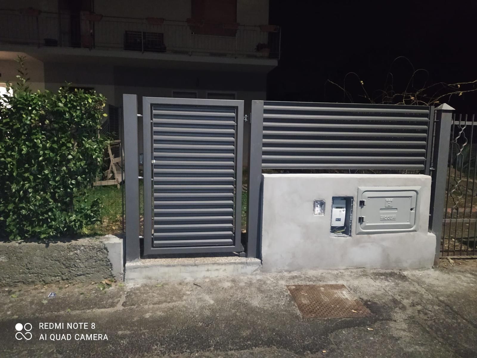 Cancello e recinzione in metallo grigio con scatole di derivazione incastonate in un muro di cemento accanto a una casa, di notte.