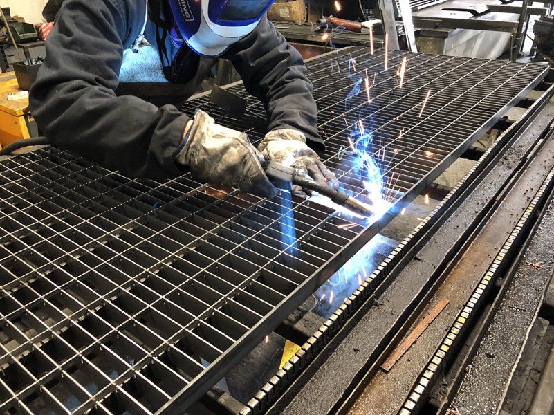 Un operaio salda una grata metallica, con scintille blu brillanti, in un'officina industriale.