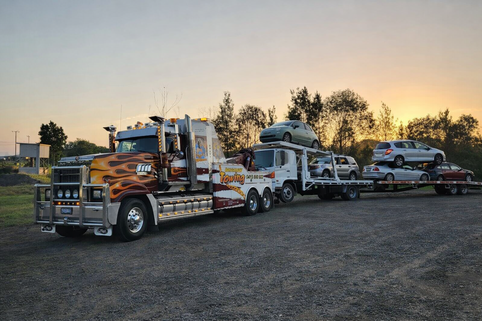 Tow truck hauling multiple cars on a flatbed trailer at sunset in an open lot — Bangalow Towing in Bangalow, NSW