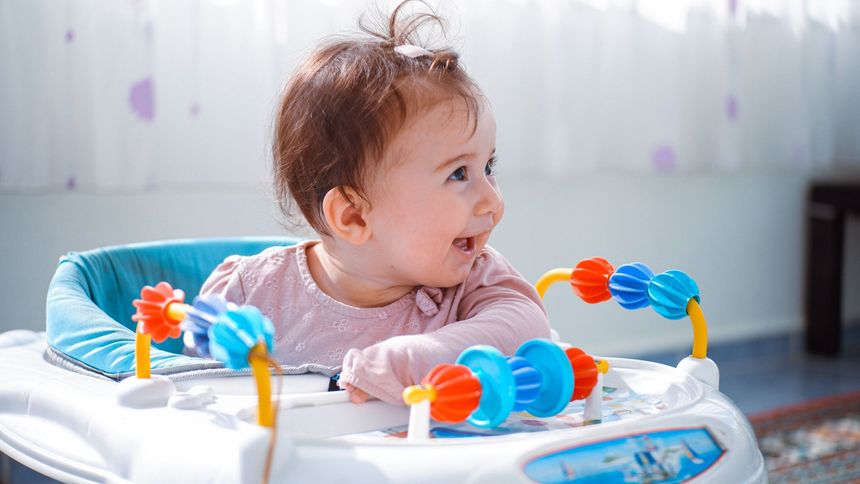 Baby lying in a white activity center, looking right, with colorful toys around them.