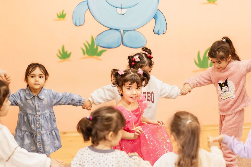 Children dancing in a playroom, holding hands in a circle with colorful wall art behind them