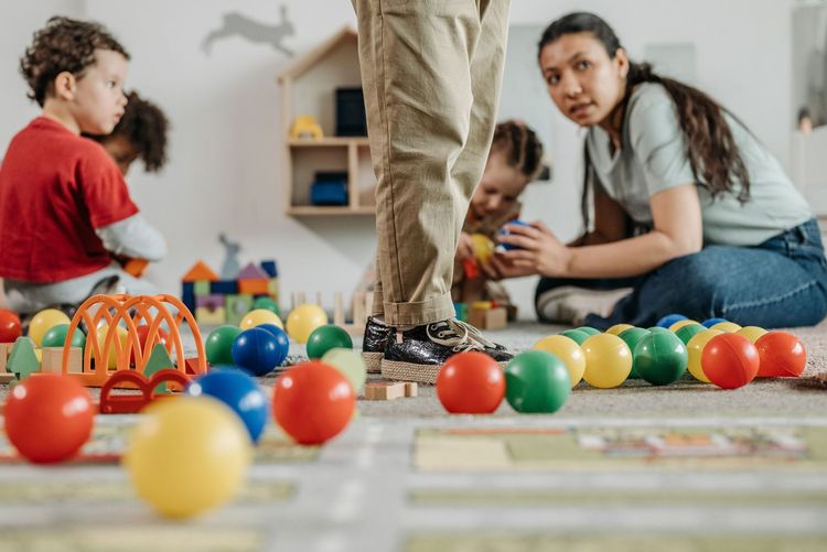 Children playing with colorful balls on a floor while an adult stands nearby in a playroom