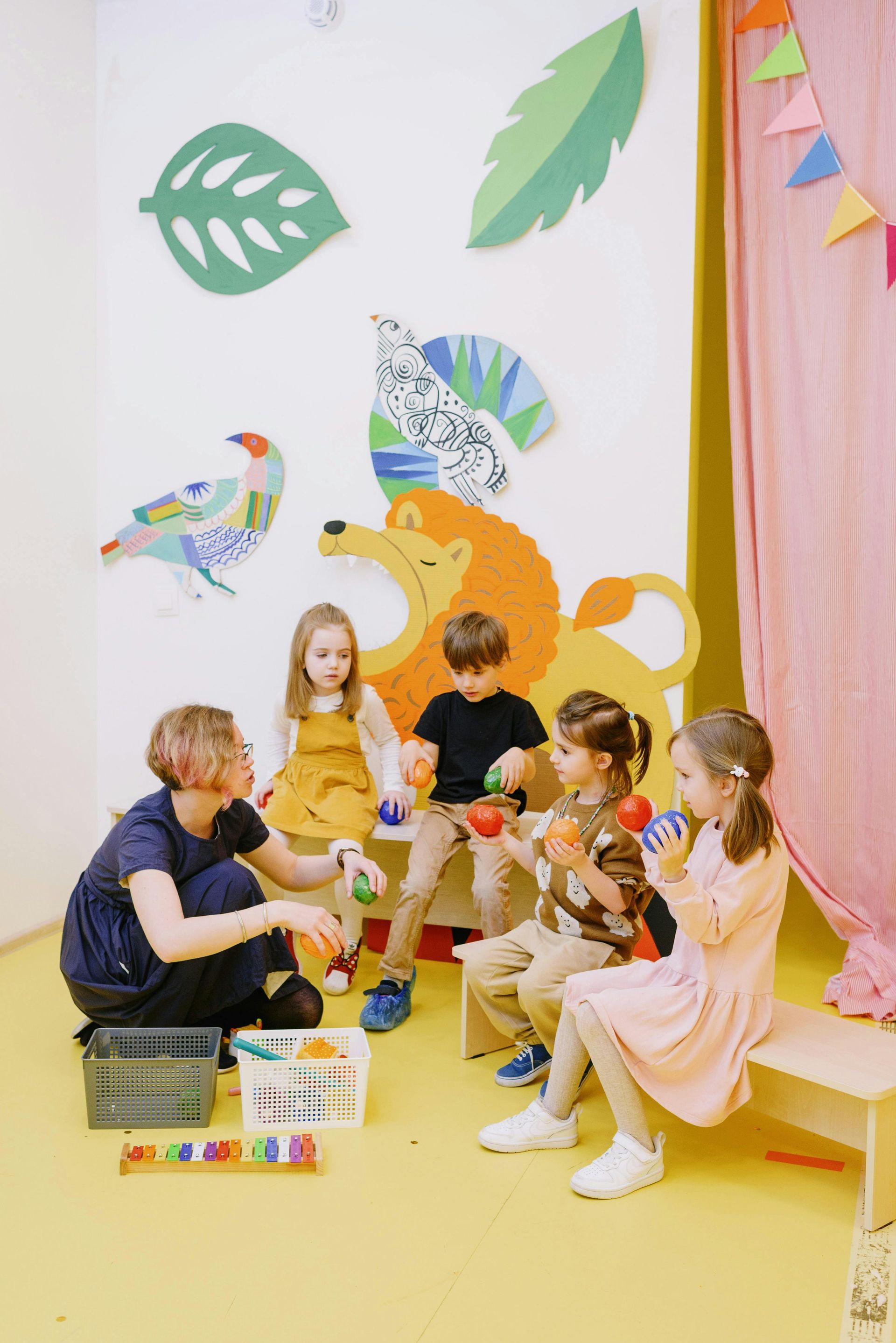 Children playing in a bright classroom with a teacher, colorful bird decorations, and toys on the floor
