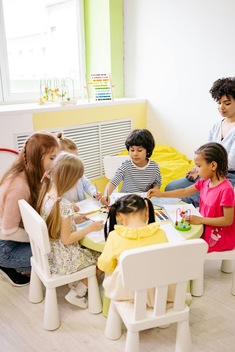 Children gathered around a table in a bright room, playing with toys and crafts.