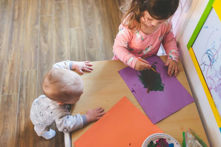 Two children painting on colored paper at a table, with a paint palette nearby.