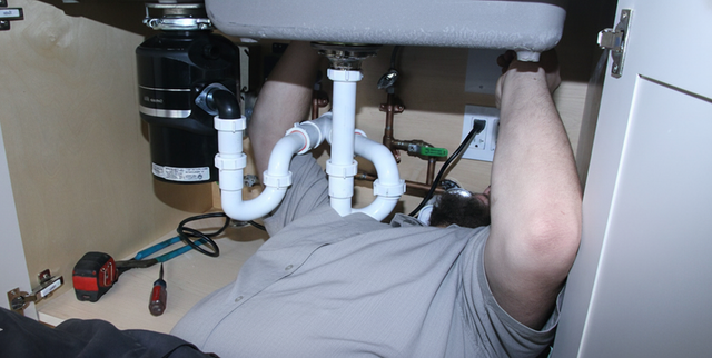 A person working under a kitchen sink, surrounded by plumbing and tools.