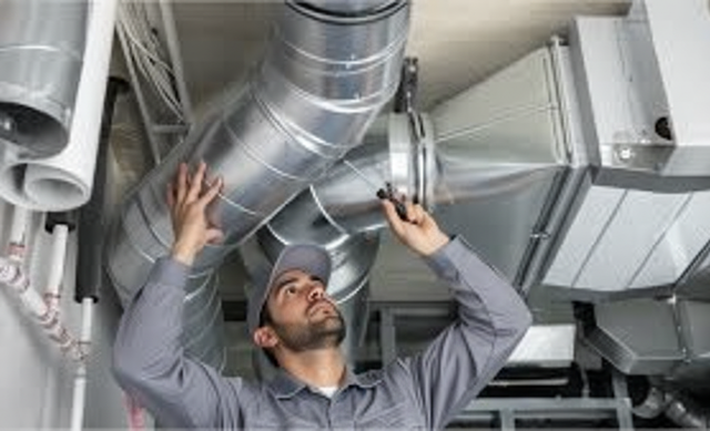 HVAC technician inspecting ventilation ducts. Indoors, looking up, holding a tool. Metal ducts and gray uniform.