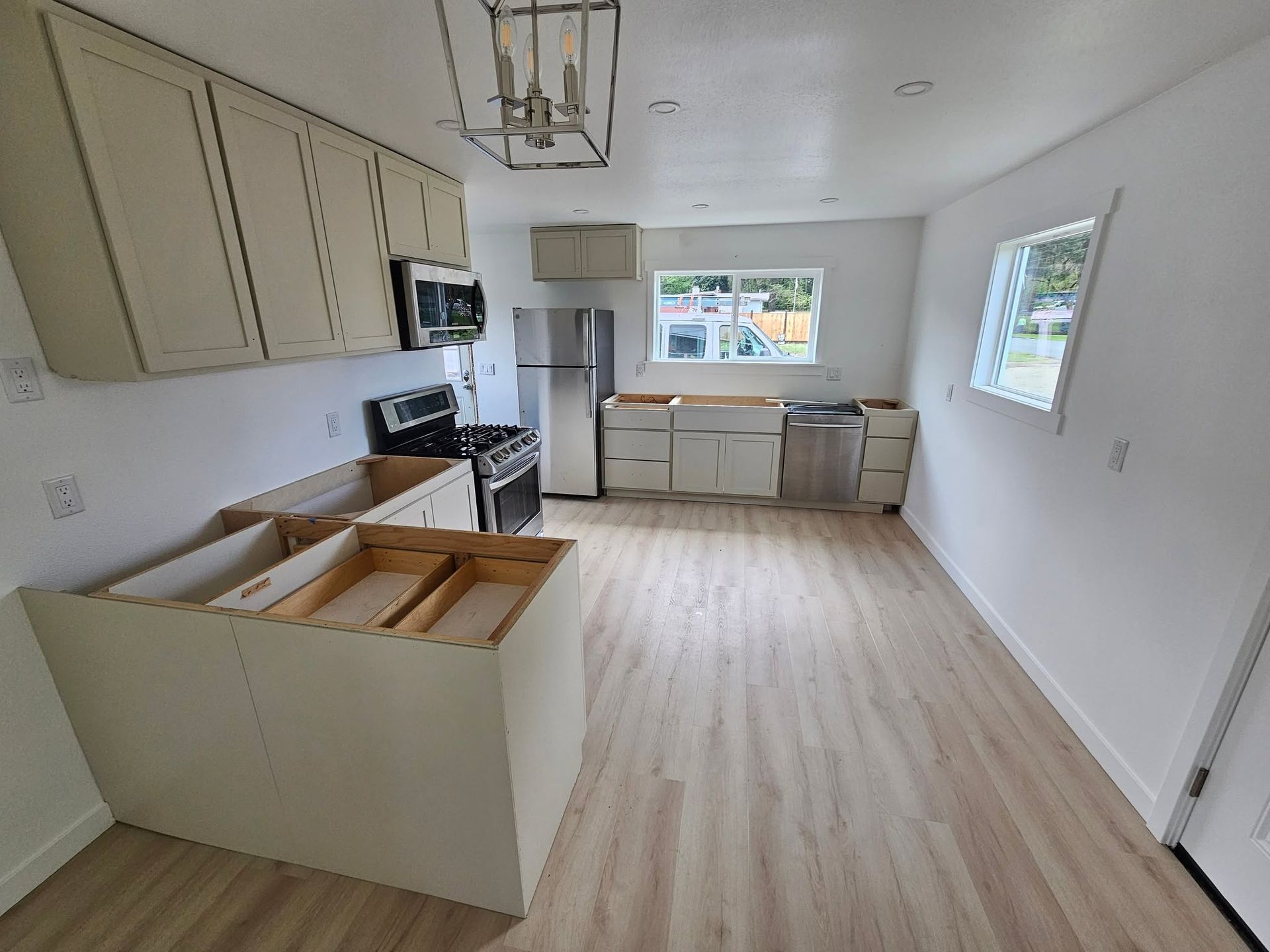 Bright empty kitchen with white cabinets, light wood floors, and windows at the far end