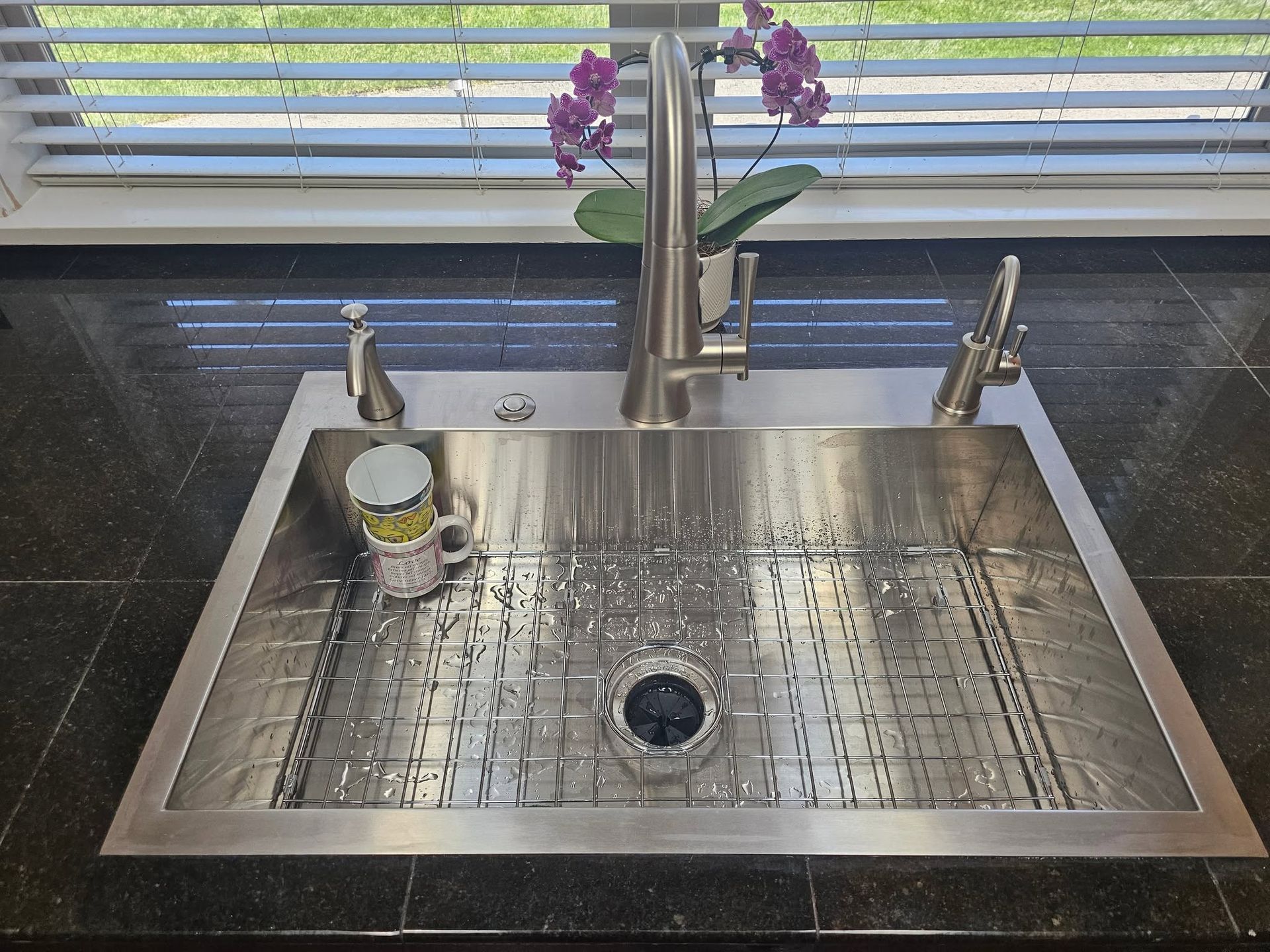 Stainless steel kitchen sink under a window with purple flowers and a soap dispenser on the left.