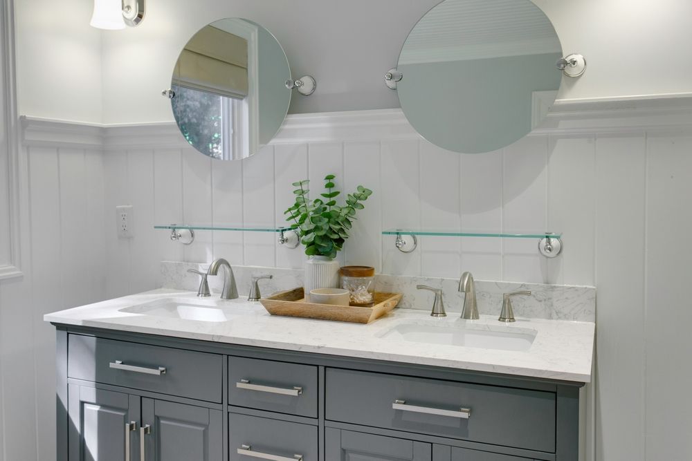 Gray double sink vanity with white countertop, round mirrors, and greenery in a white bathroom. — Dimond J Carpentry in Bli Bli, QLD