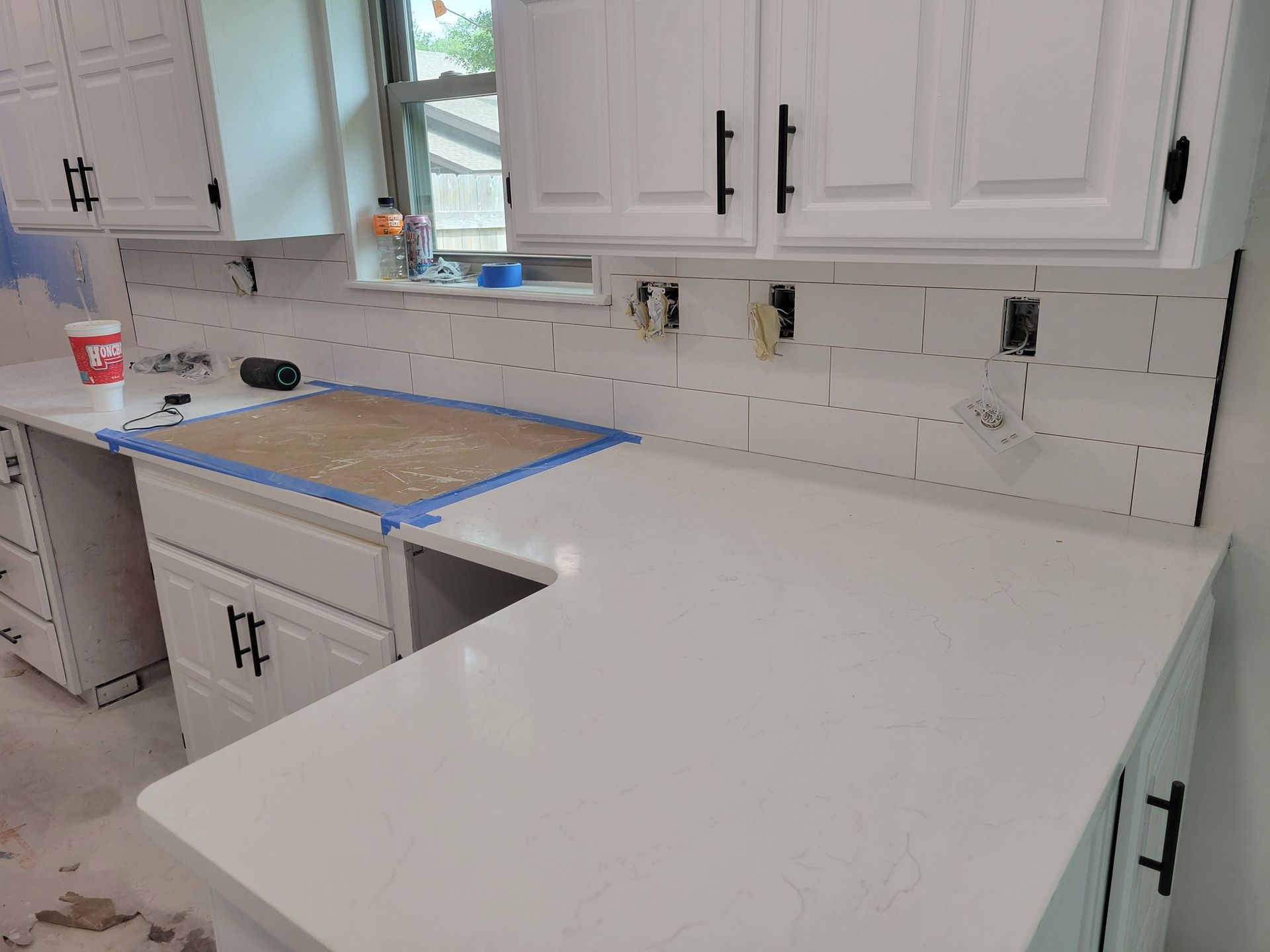 White kitchen with new L-shaped countertop installed, sink cutout taped off, and cabinets unfinished around the edges