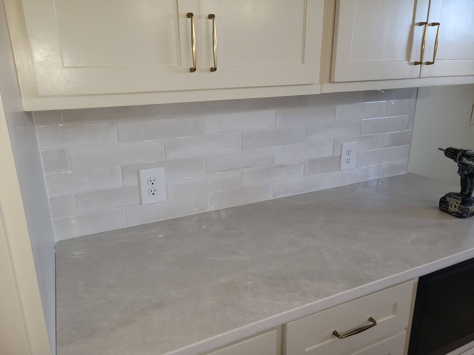 Kitchen countertop with white cabinets, light gray tile backsplash, and a small drill on the right.