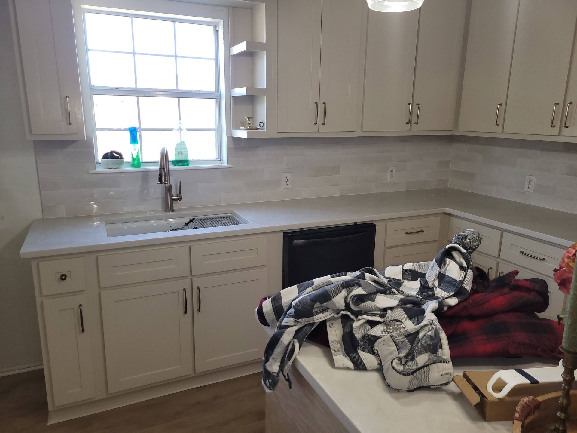 Messy kitchen with beige cabinets, sink by a window, and a pile of clothes on the counter and table.
