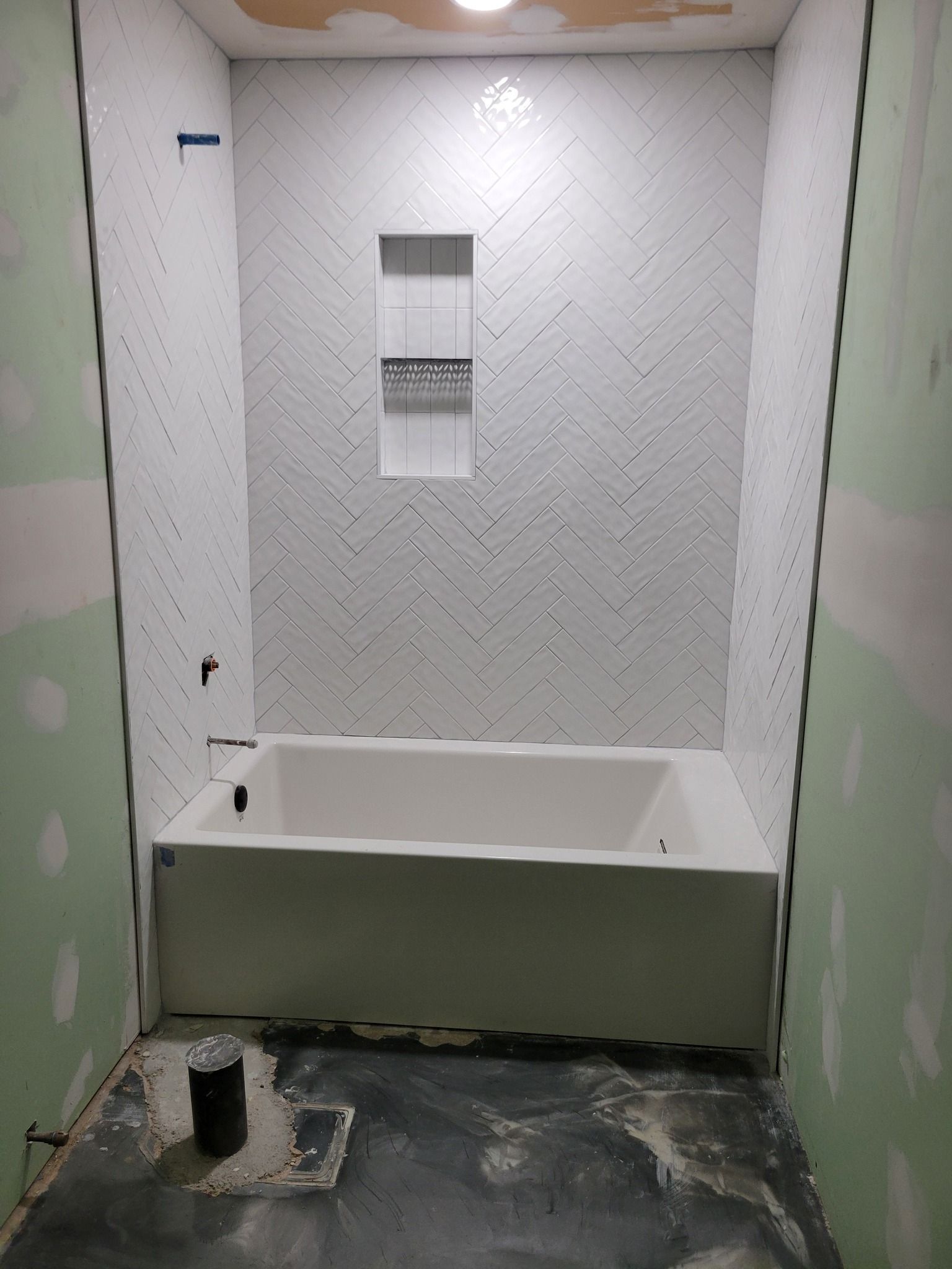 Partially tiled bathroom with a white bathtub, unfinished green walls, and a floor drain in front