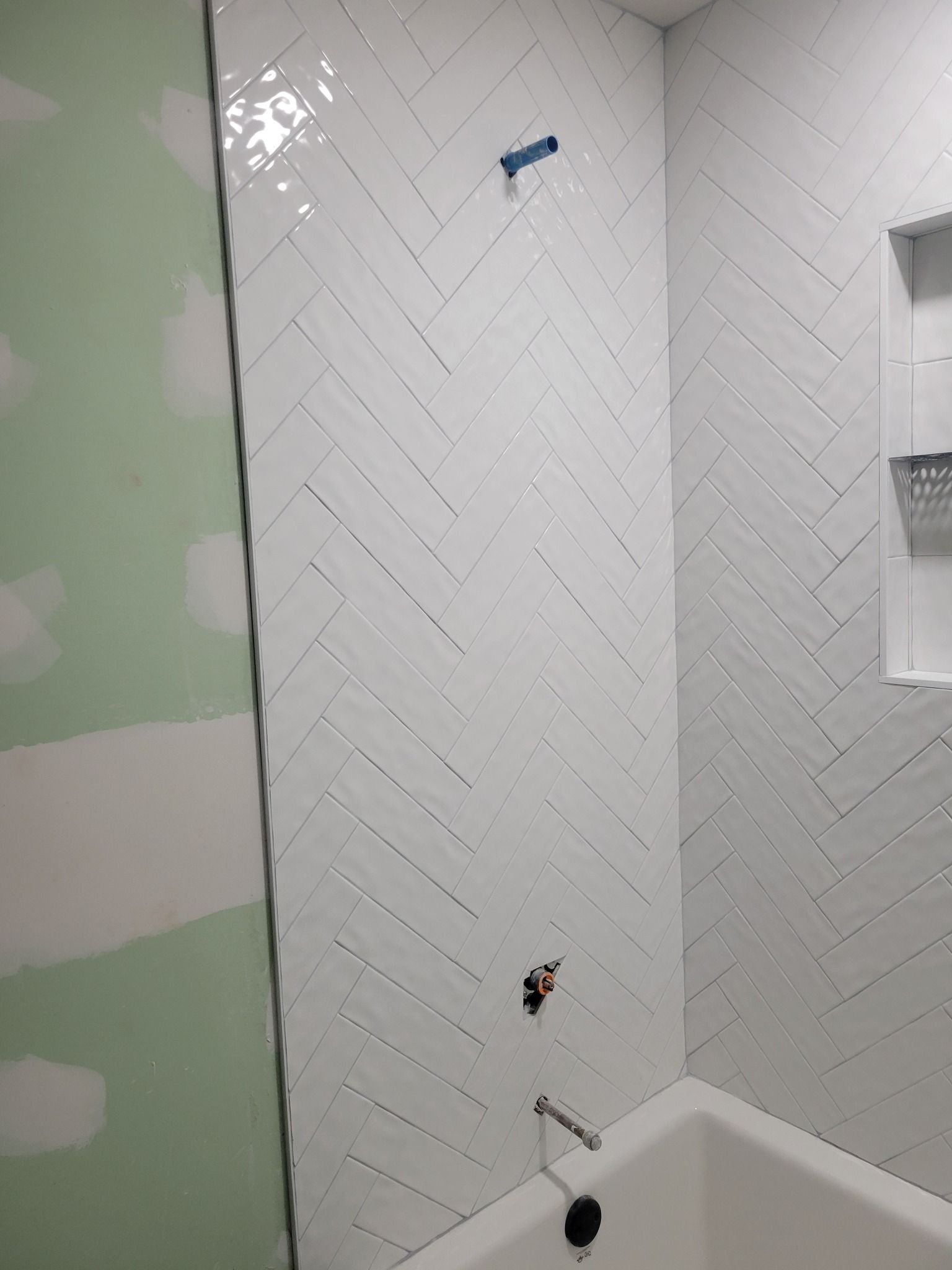 Bathroom wall with white herringbone tile, a tub corner, and green unfinished drywall at left