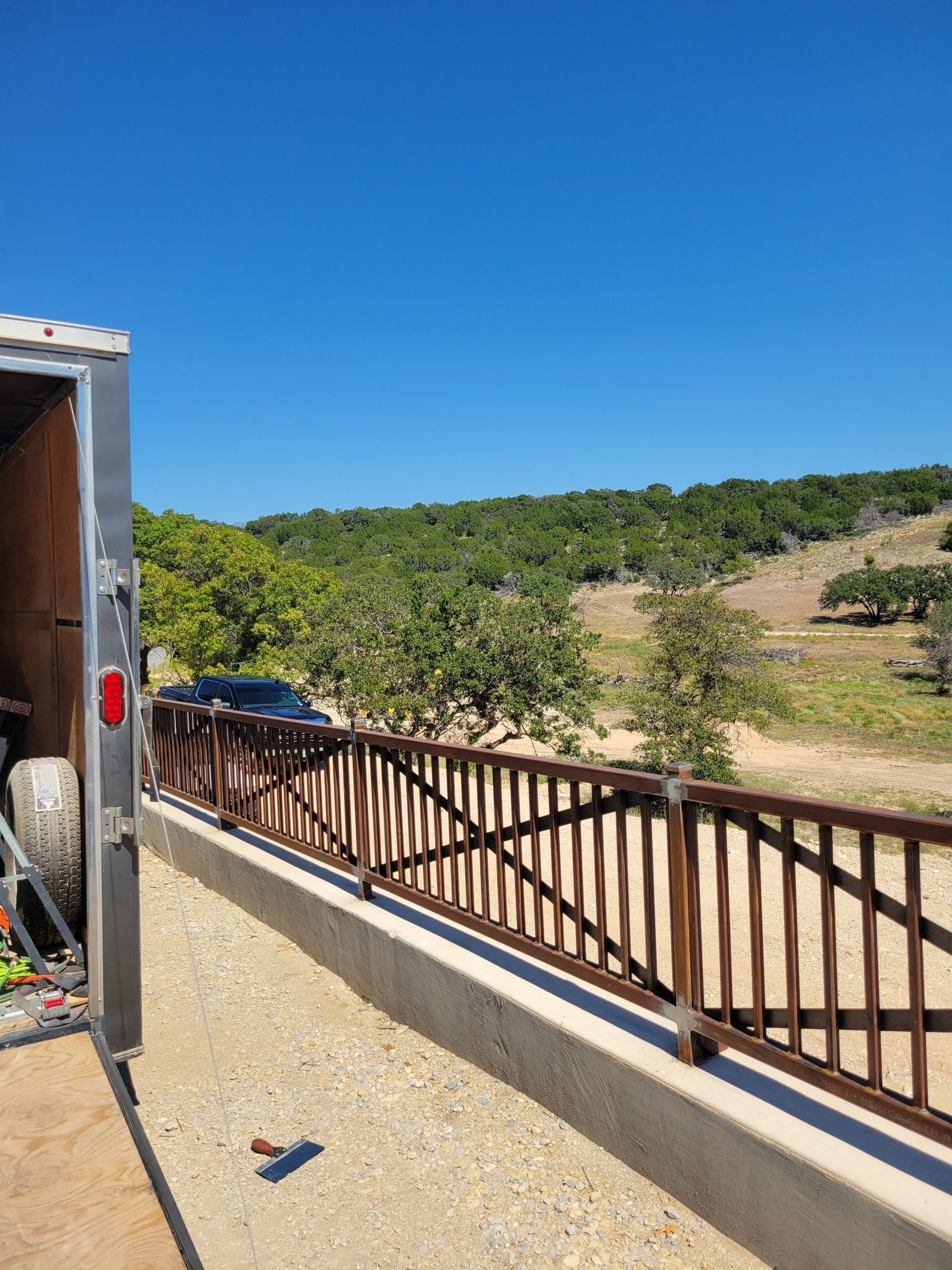 Outdoor balcony with wooden railing overlooking a sunny hillside and blue sky, with a truck partially visible on the left.