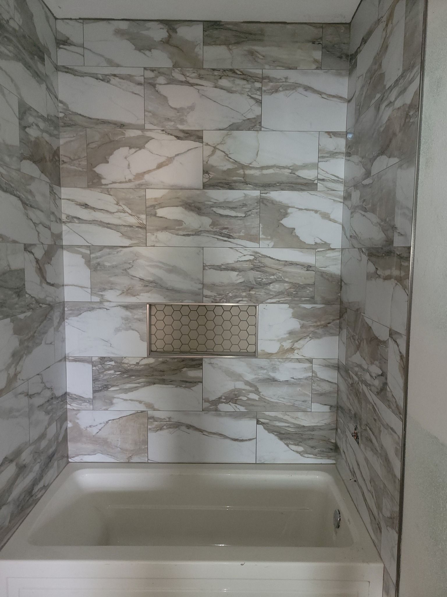 Bathroom shower with marble-pattern tile walls, built-in niches, and a bathtub below.