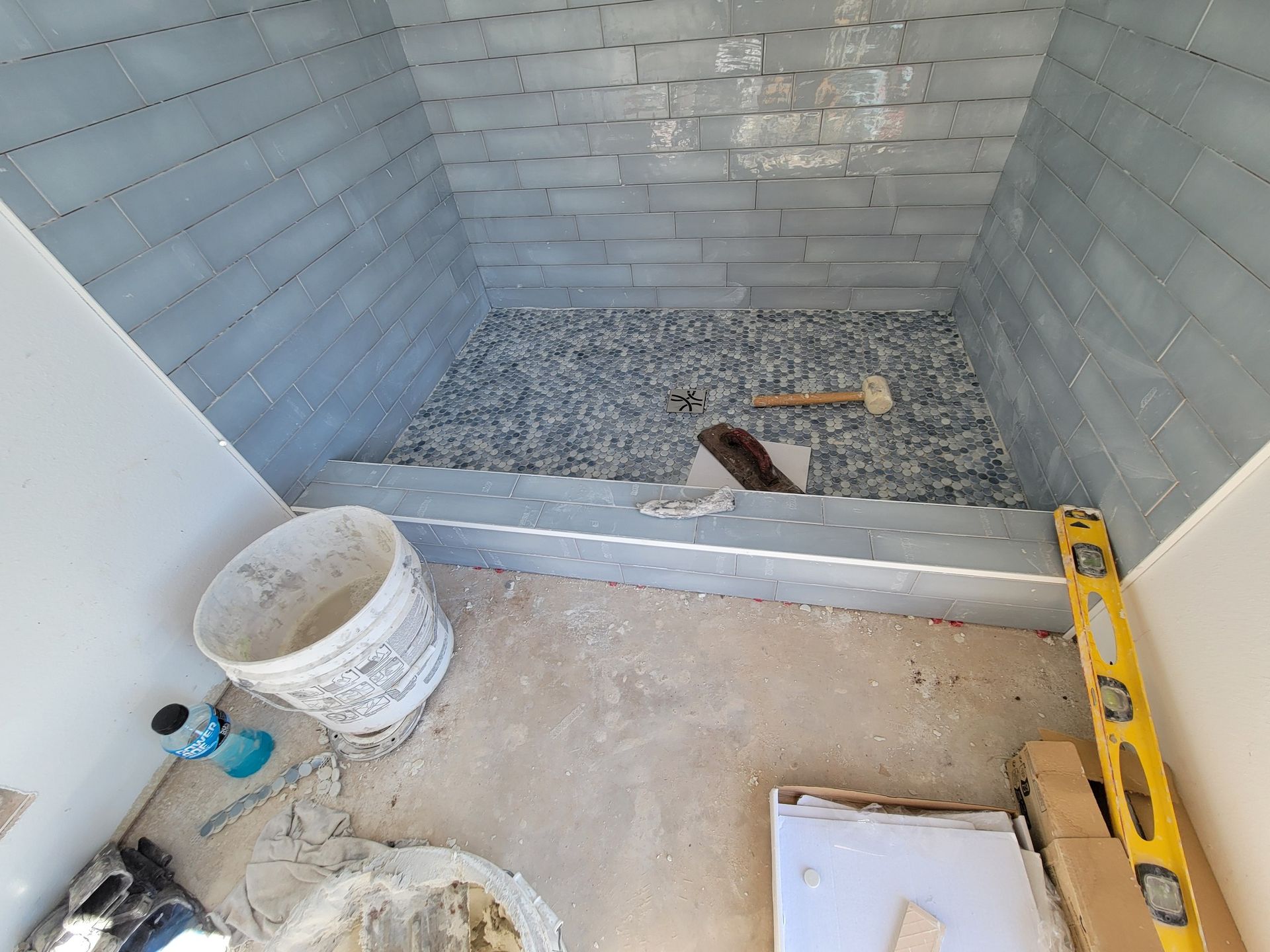 Small unfinished shower stall with gray tile, gravel floor, bucket, and level on the concrete outside area