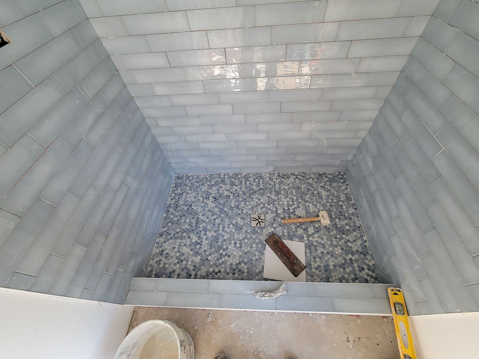 Small tiled shower stall with gravel floor, a drain opening, and a wooden tool inside.
