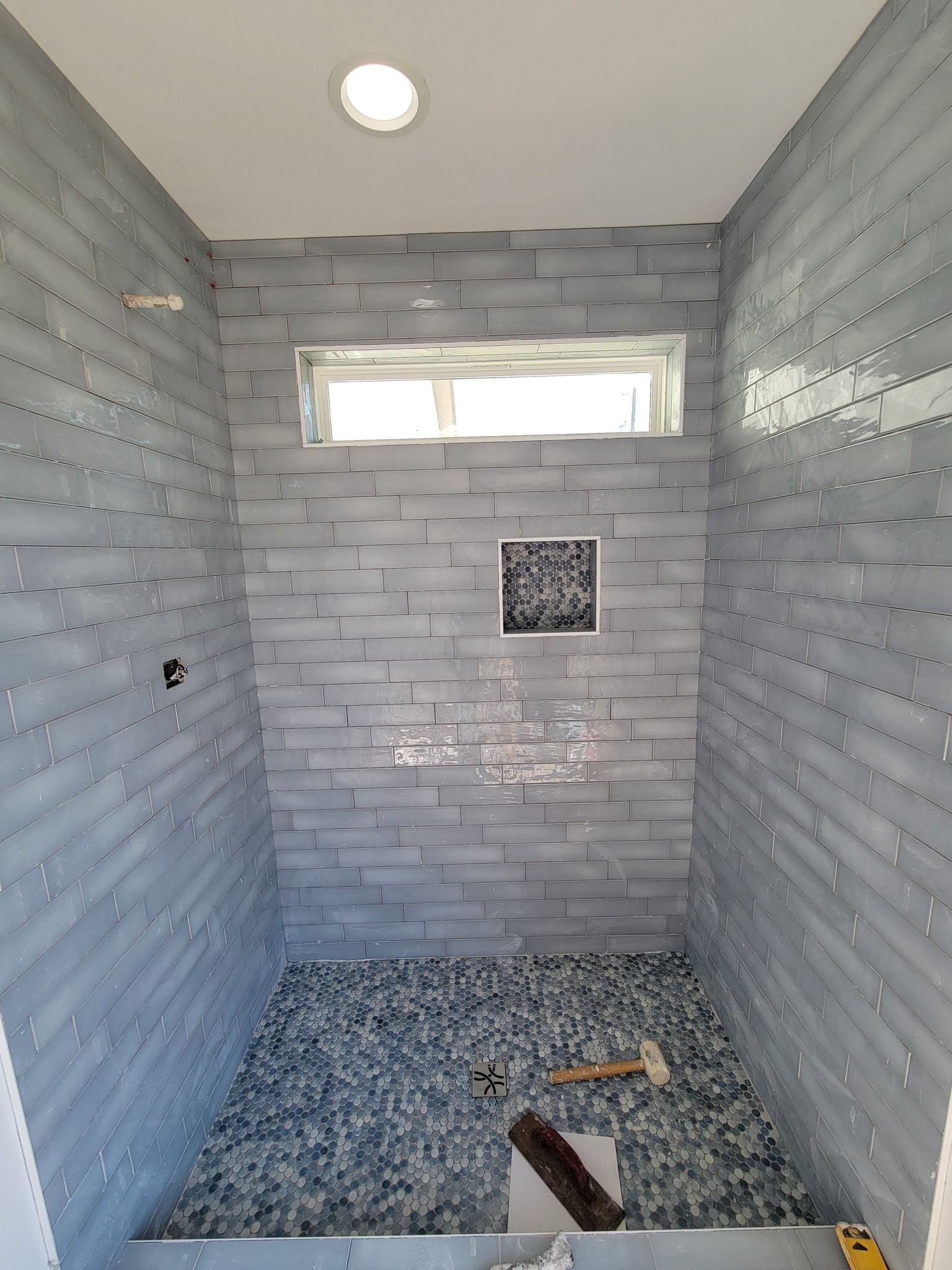 Empty tiled shower stall with a small window and floor drain, gray walls and pebble floor.