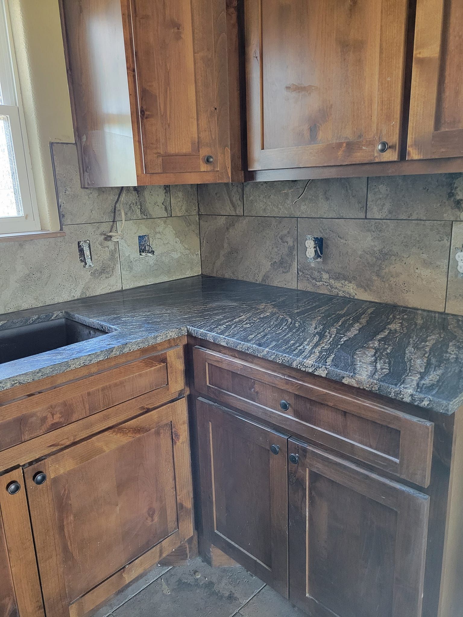 Corner kitchen with wood cabinets, dark granite countertop, stone tile backsplash, and a sink by the window