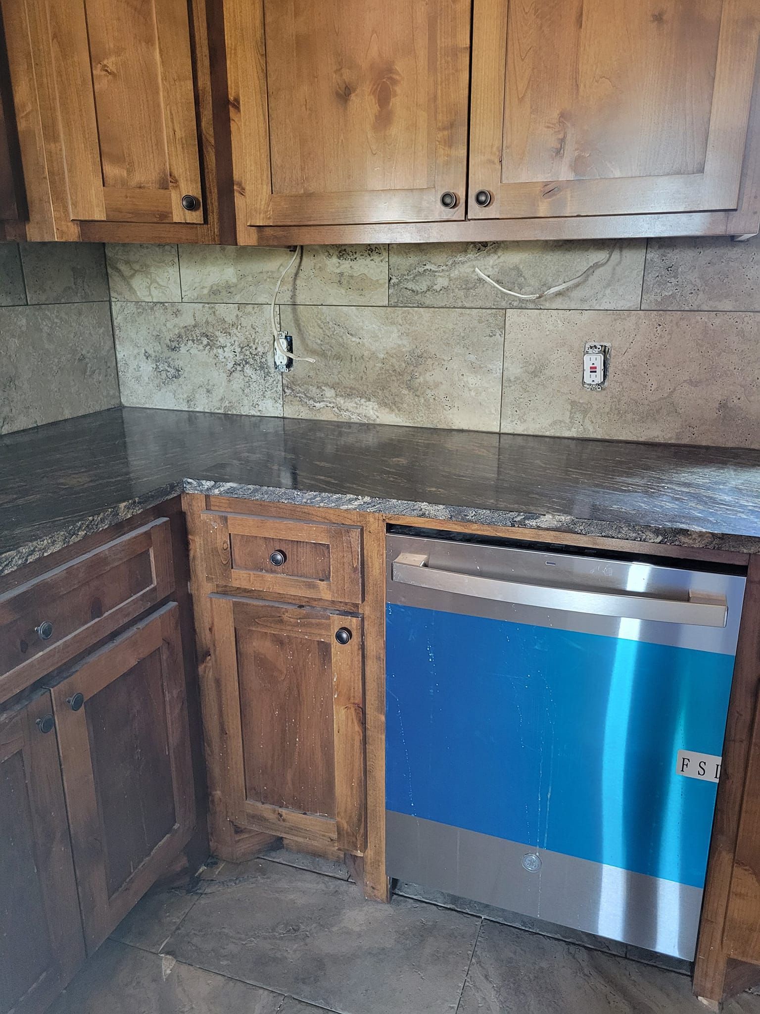 Kitchen with wooden cabinets, dark countertop, stone backsplash, and a blue-and-silver dishwasher below the counter.