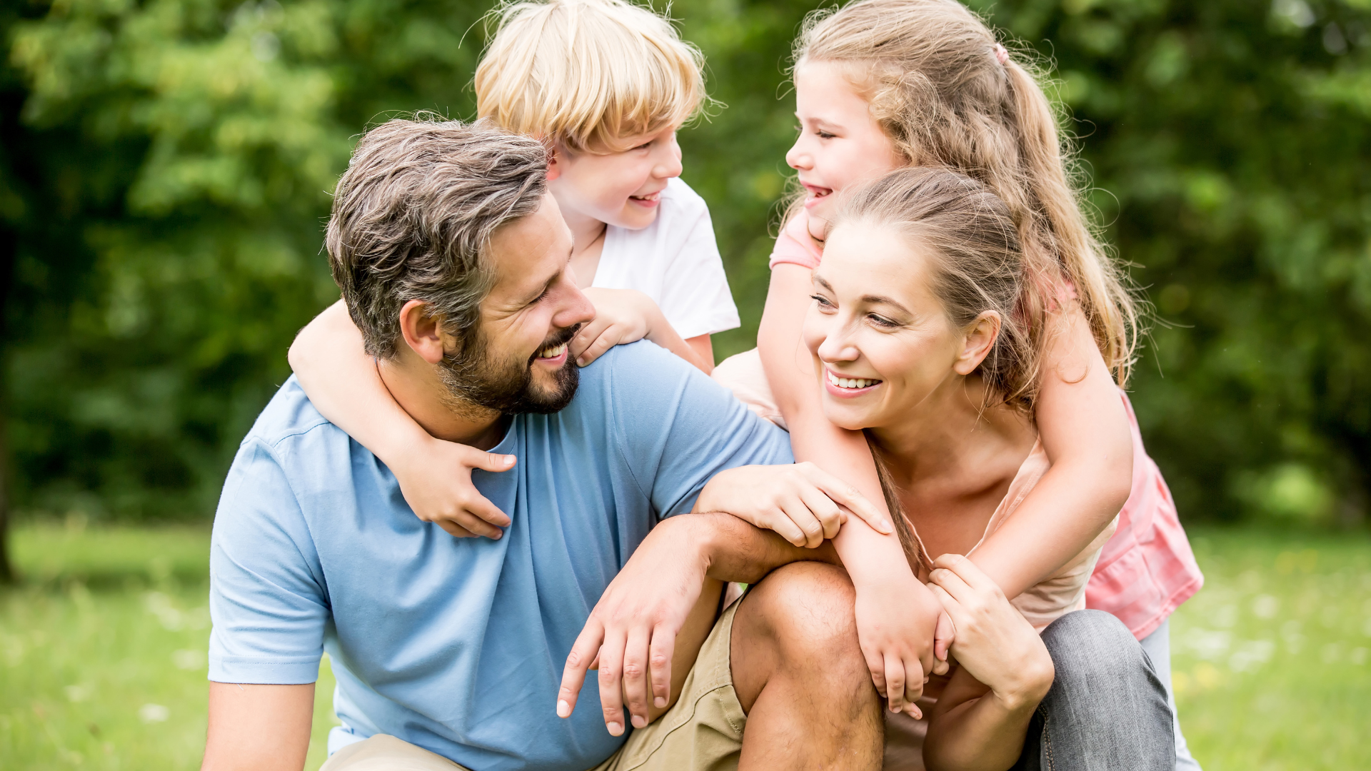 Family of four smiling, hugging, and enjoying time outdoors in a park.