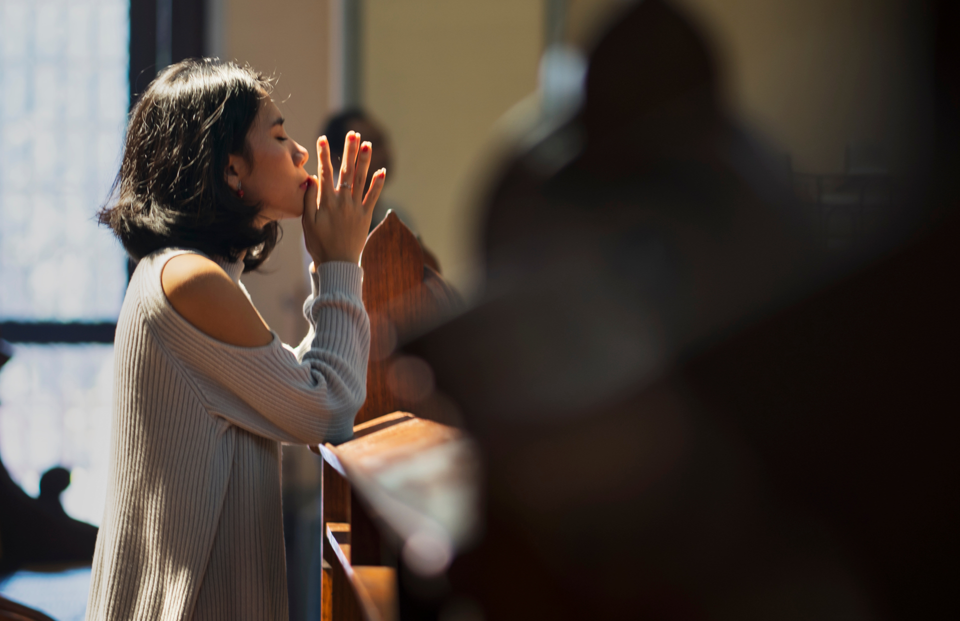 Woman with hands clasped in prayer, eyes closed in church, sunlight streaming in.