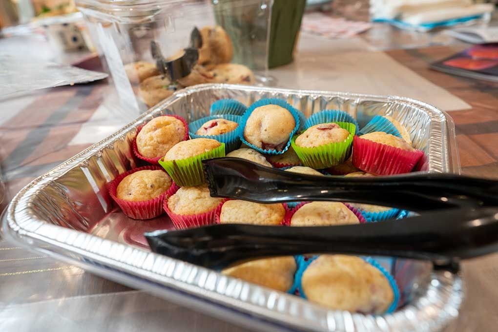 Mini muffins in colorful paper cups in a foil container with black serving tongs.