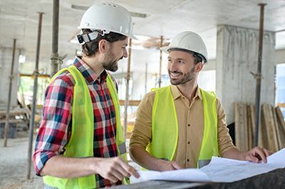 Two construction workers in hard hats and vests examining blueprints in a building under construction.