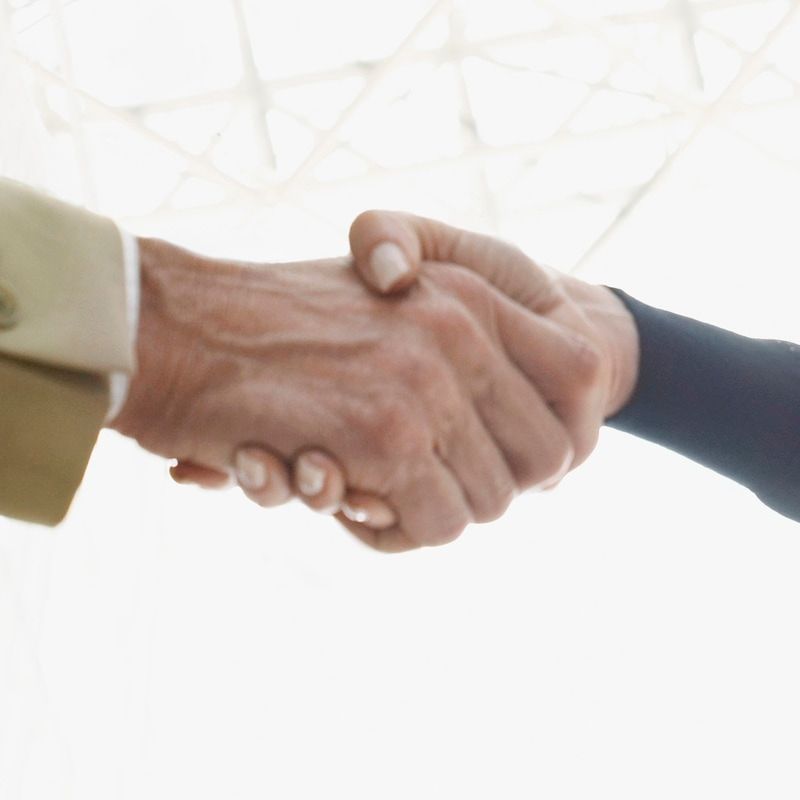 Two hands clasped in a handshake. One hand has a tan suit jacket sleeve, the other a black sleeve.