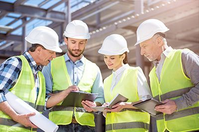 Construction workers in white hard hats and safety vests reviewing plans on a construction site.