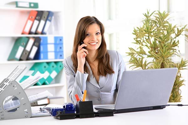 Woman in a gray suit smiles while talking on a phone, sitting at a desk with a laptop and office supplies.