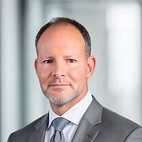 Headshot of a man in a gray suit and tie with a neutral expression against a blurred office background