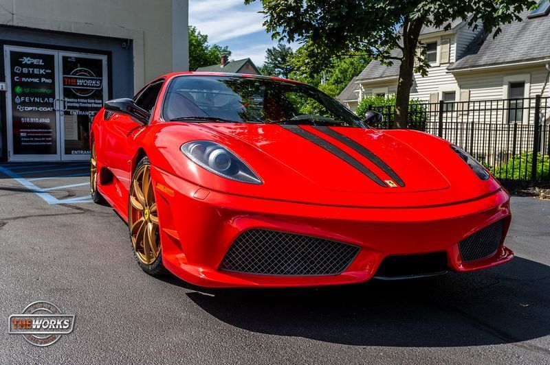 A red Ferrari F430 with racing stripes and gold wheels parked on a driveway in front of a building.