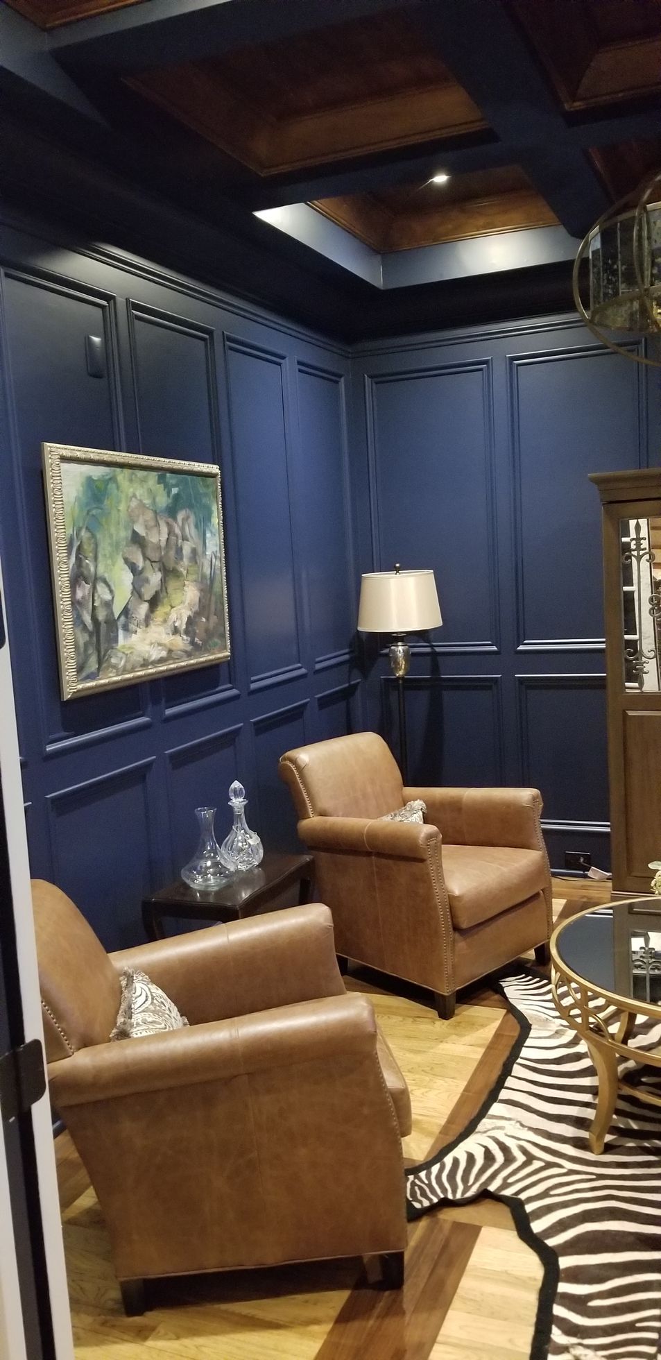 Blue-paneled sitting room with leather chairs, lamp, artwork, and zebra rug under a tray ceiling