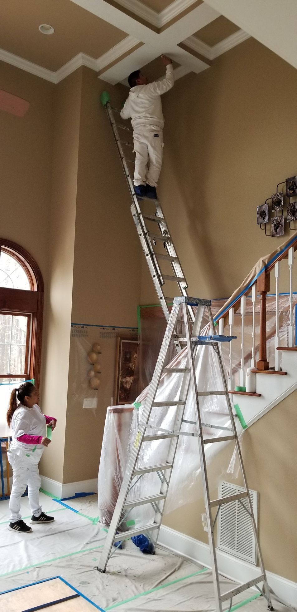 Workers on ladders painting a tall hallway ceiling and trim.