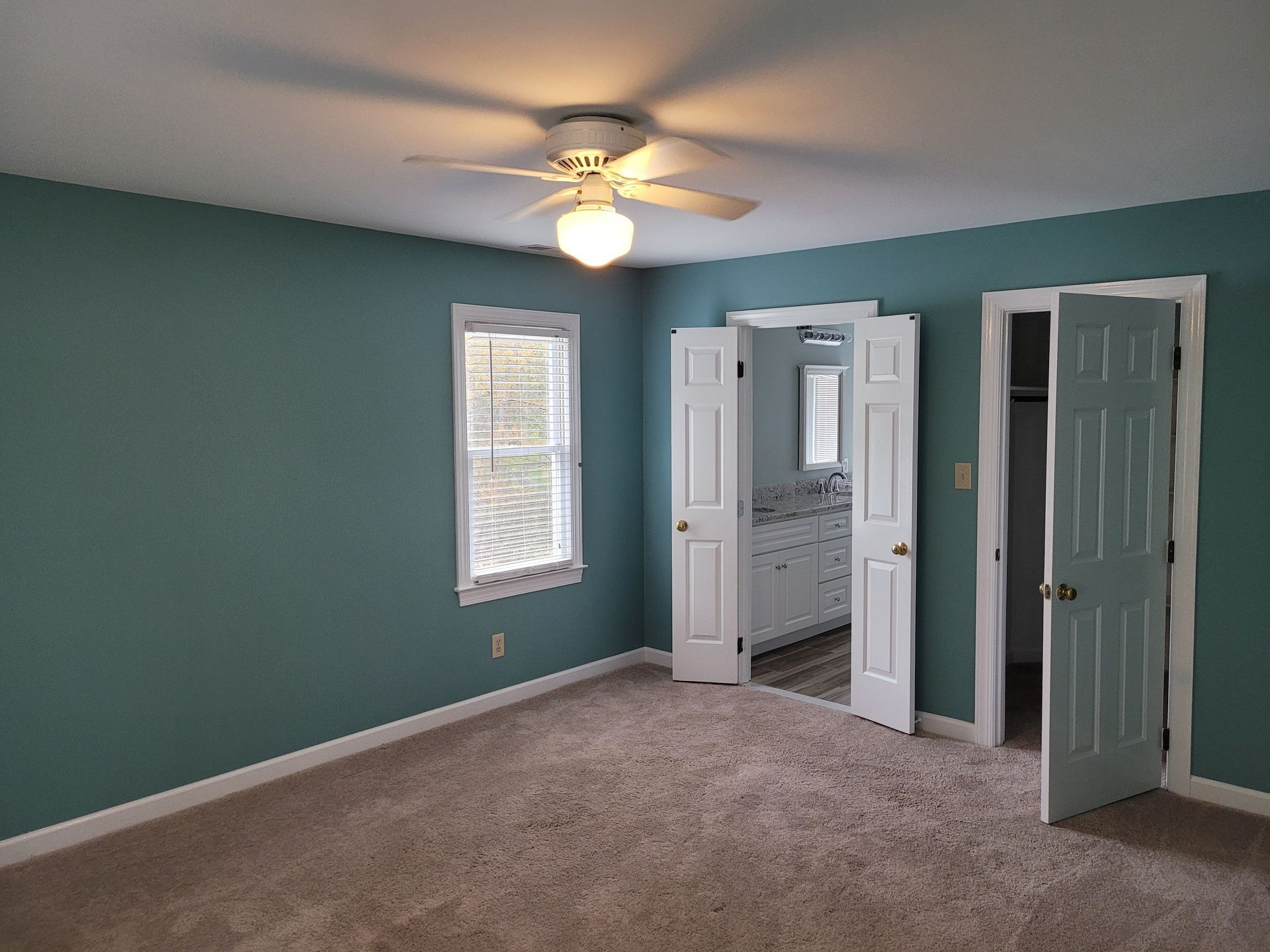 Empty teal bedroom with beige carpet, ceiling fan, window, and open closet doors.