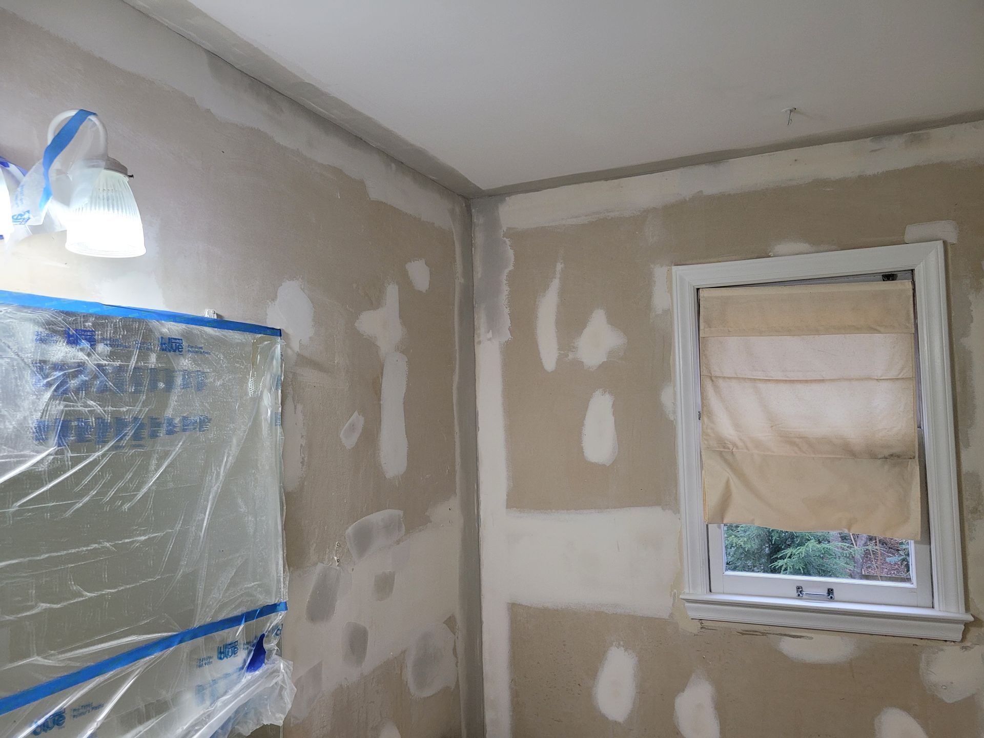 Bare room under renovation with taped plastic over a cabinet and a small window with a blind.