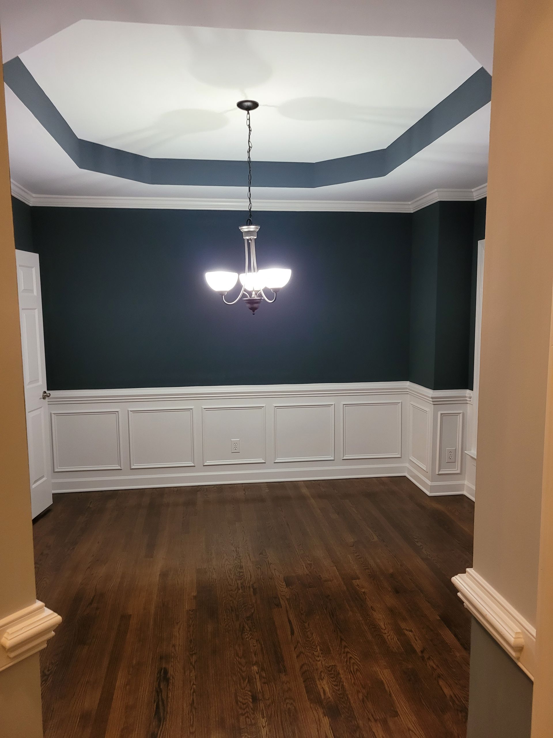 Empty dining room with dark walls, white wainscoting, hardwood floors, and a central chandelier.