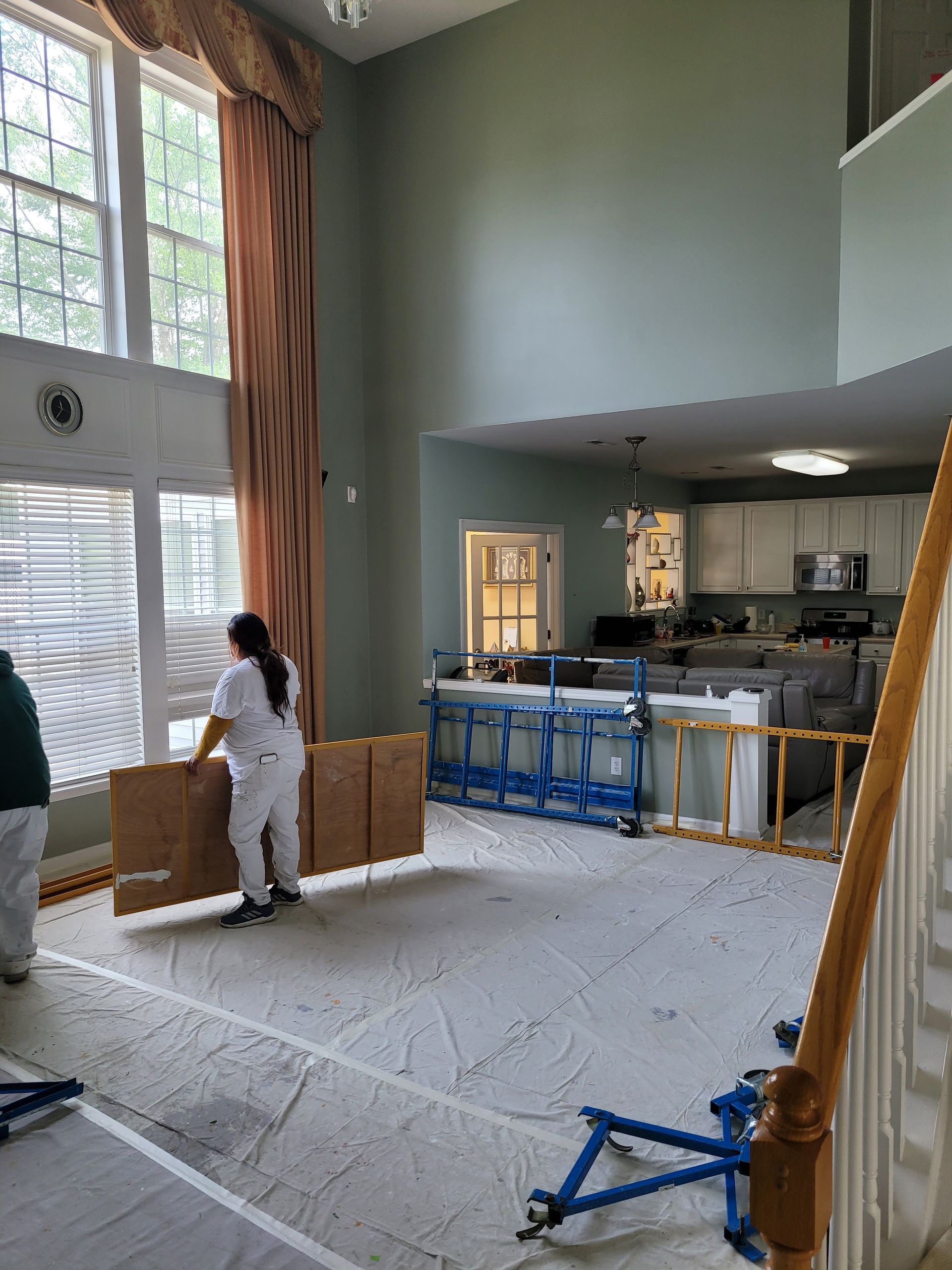 Tall living room with high windows, person moving boxes, and renovation materials on the floor.