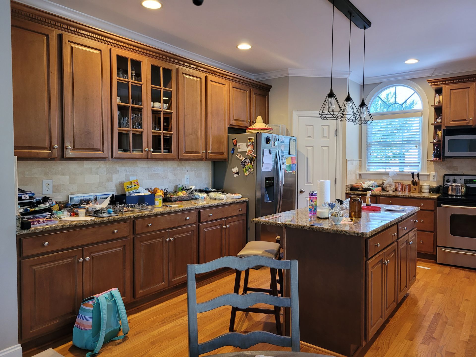 Warm kitchen with wooden cabinets, island, pendant lights, and a refrigerator by a bright window.