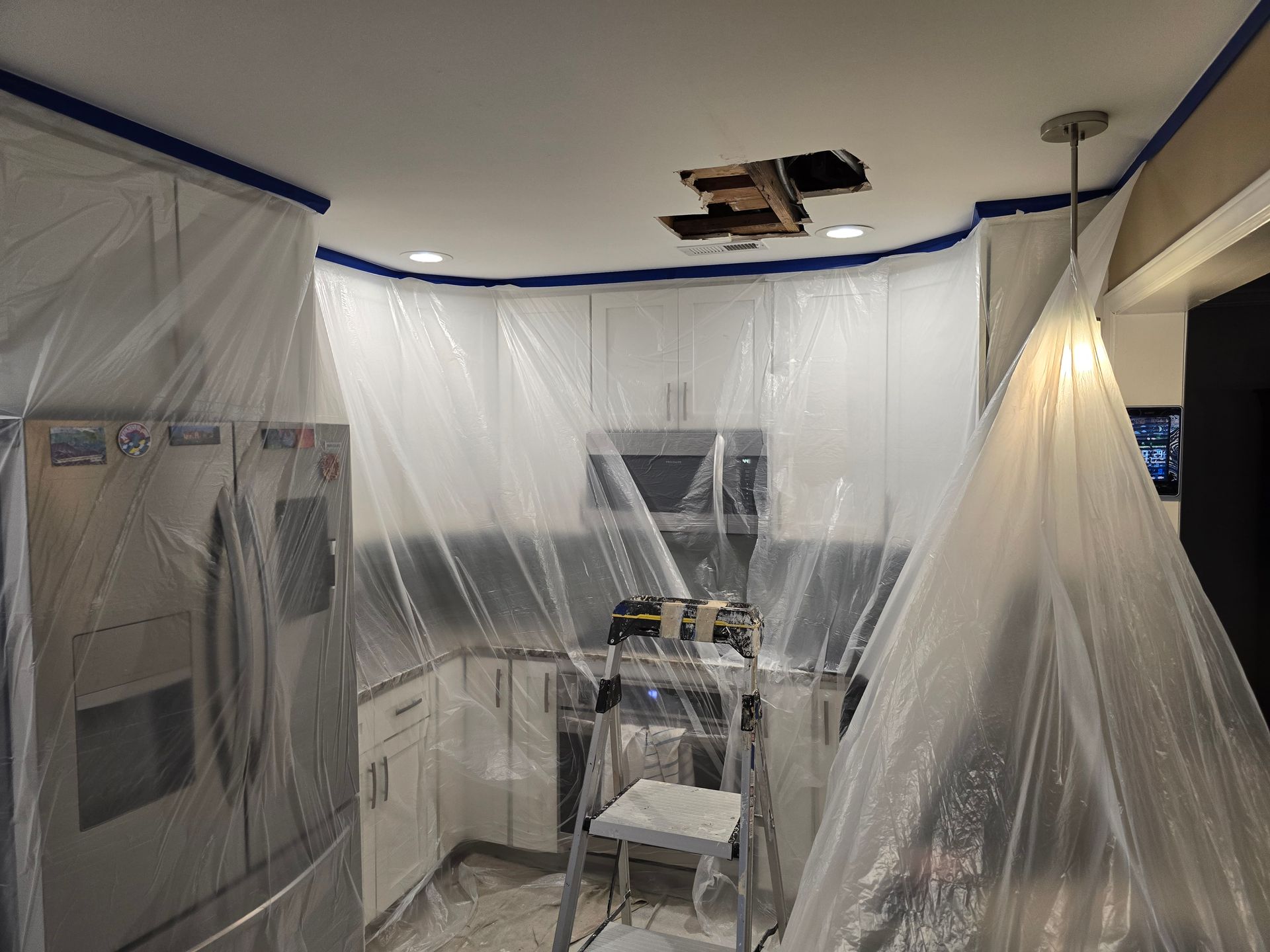 Plastic sheeting covers a kitchen under renovation, with blue painter’s tape and a stepladder in the center.