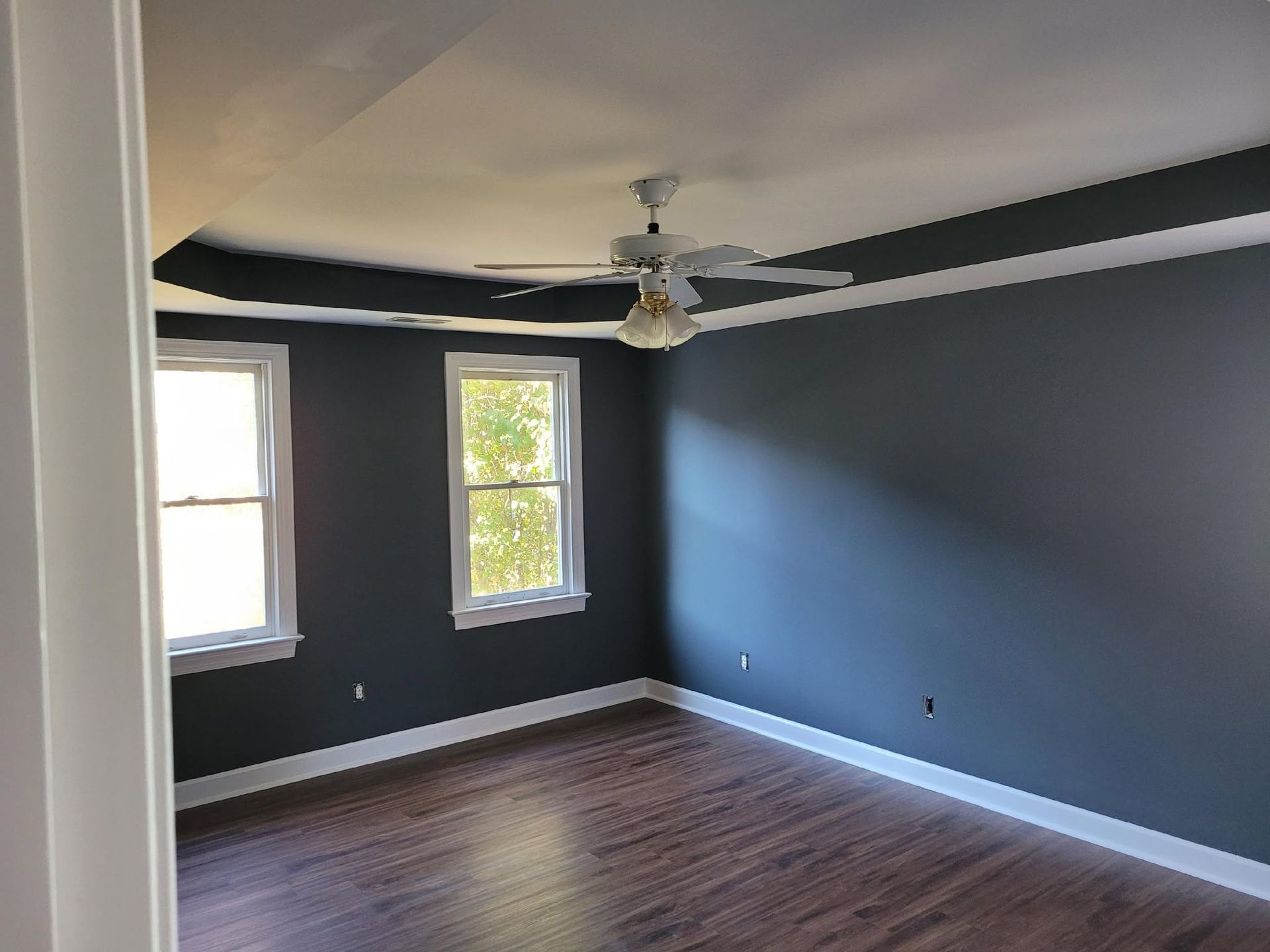 Empty dark-gray bedroom with two windows, ceiling fan, and hardwood floors.