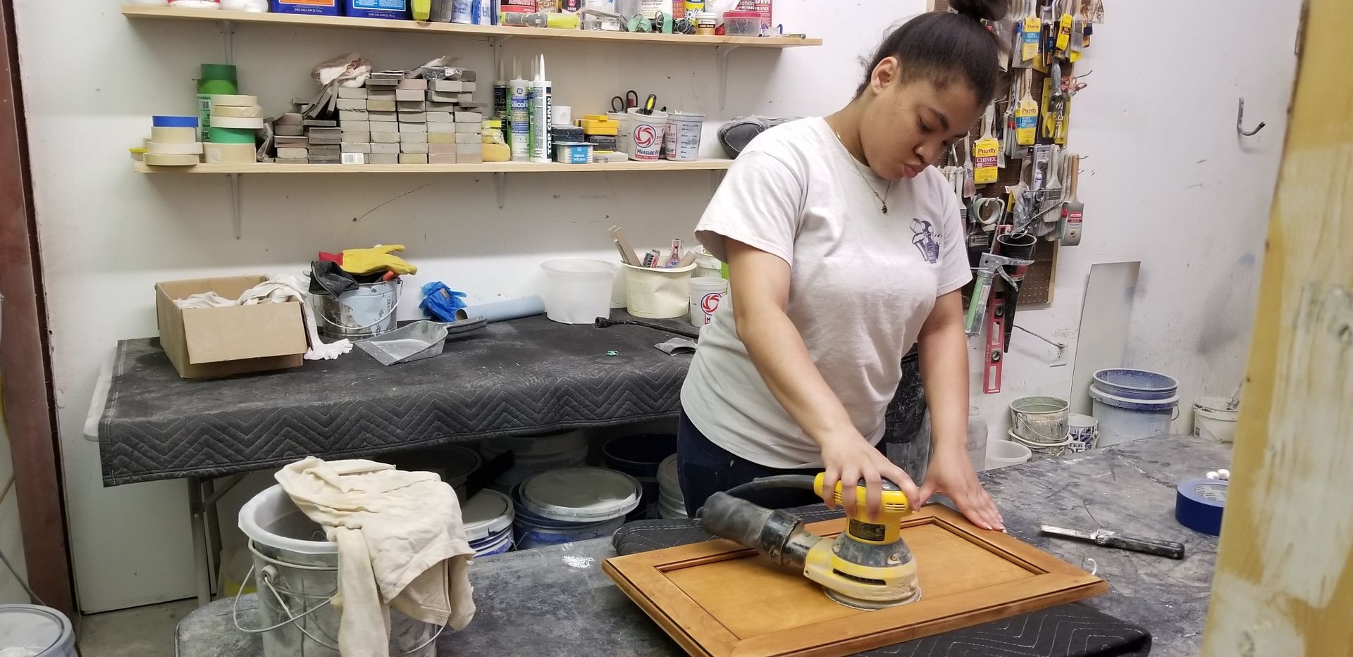 Person sanding a wooden board in a workshop, with tools and supplies on shelves behind them.