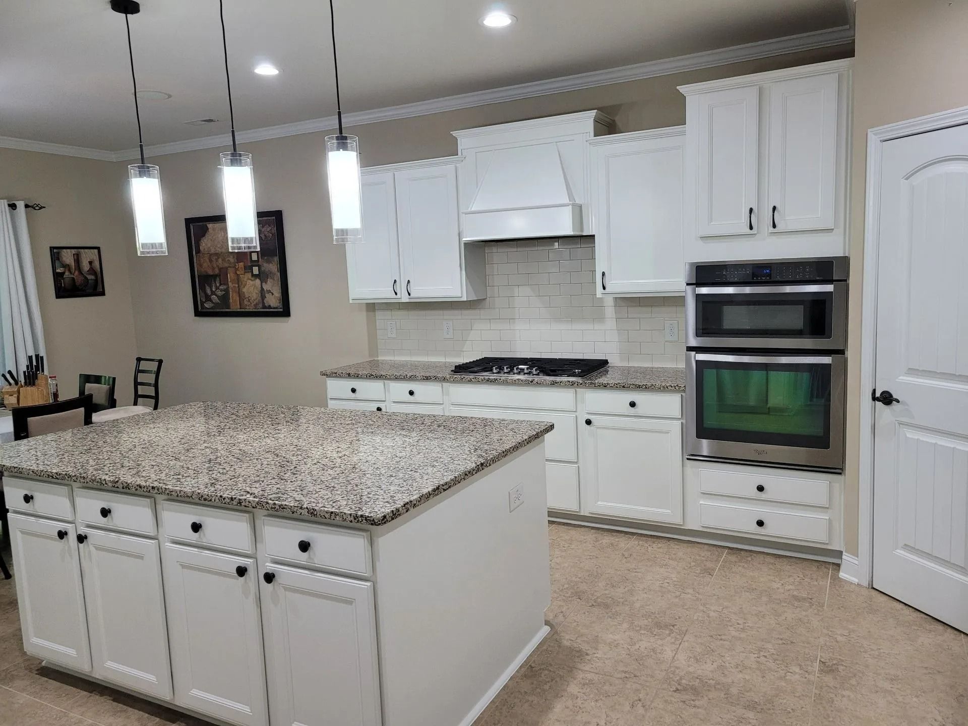 Bright kitchen with white cabinets, granite island, stainless oven, and beige tile floor.