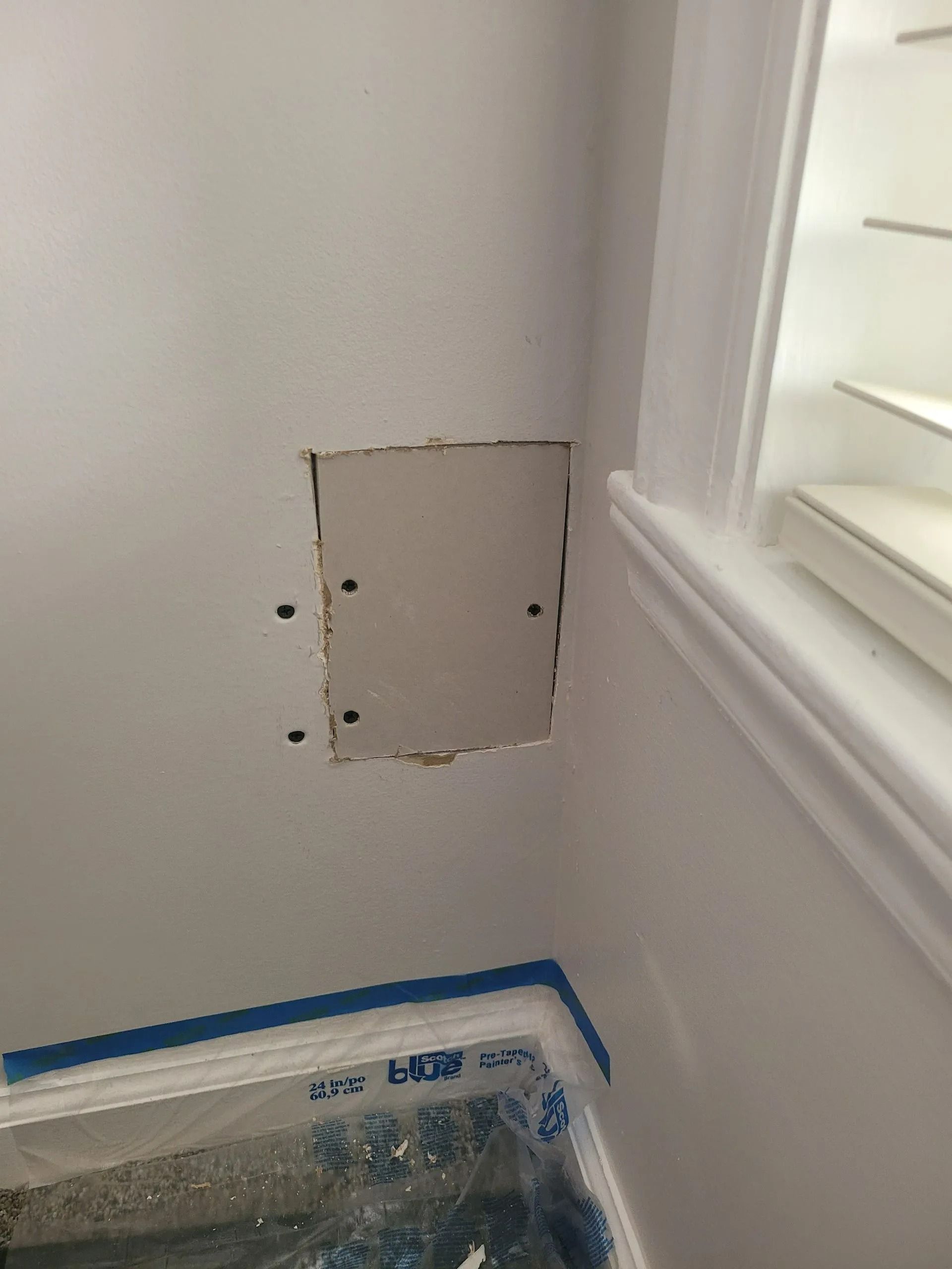 White wall with a small patched square opening beside a window, above a covered surface.